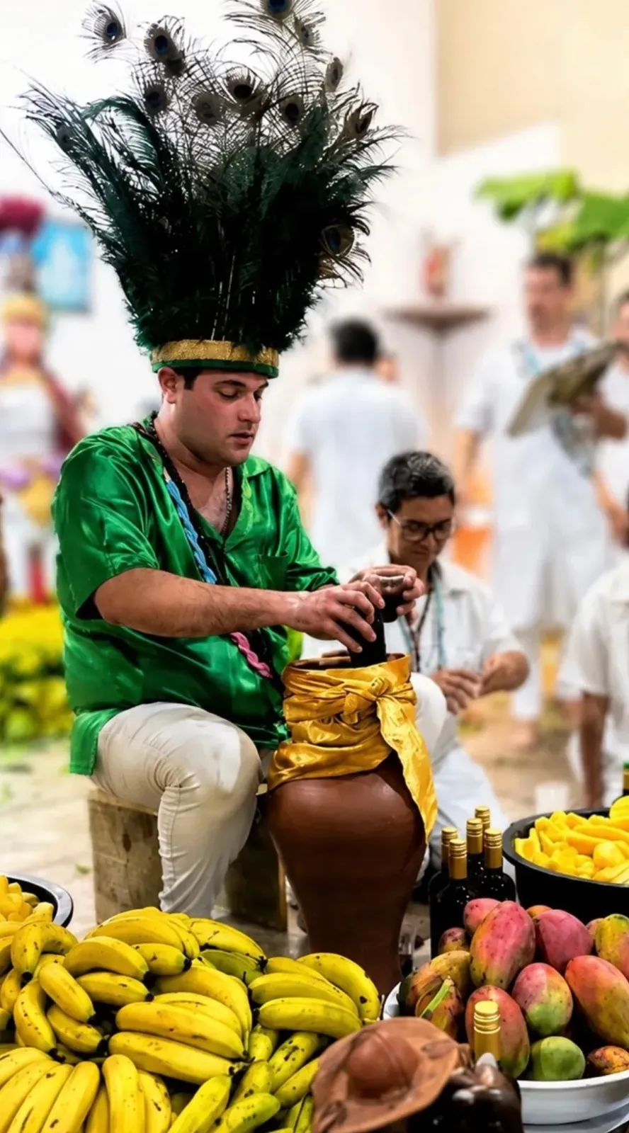Pai Luiz Fernando de cocar de penas de pavão e camisa verde, preparando a bebida dos Caboclos em um pote de barro cercado de bananas e mangas