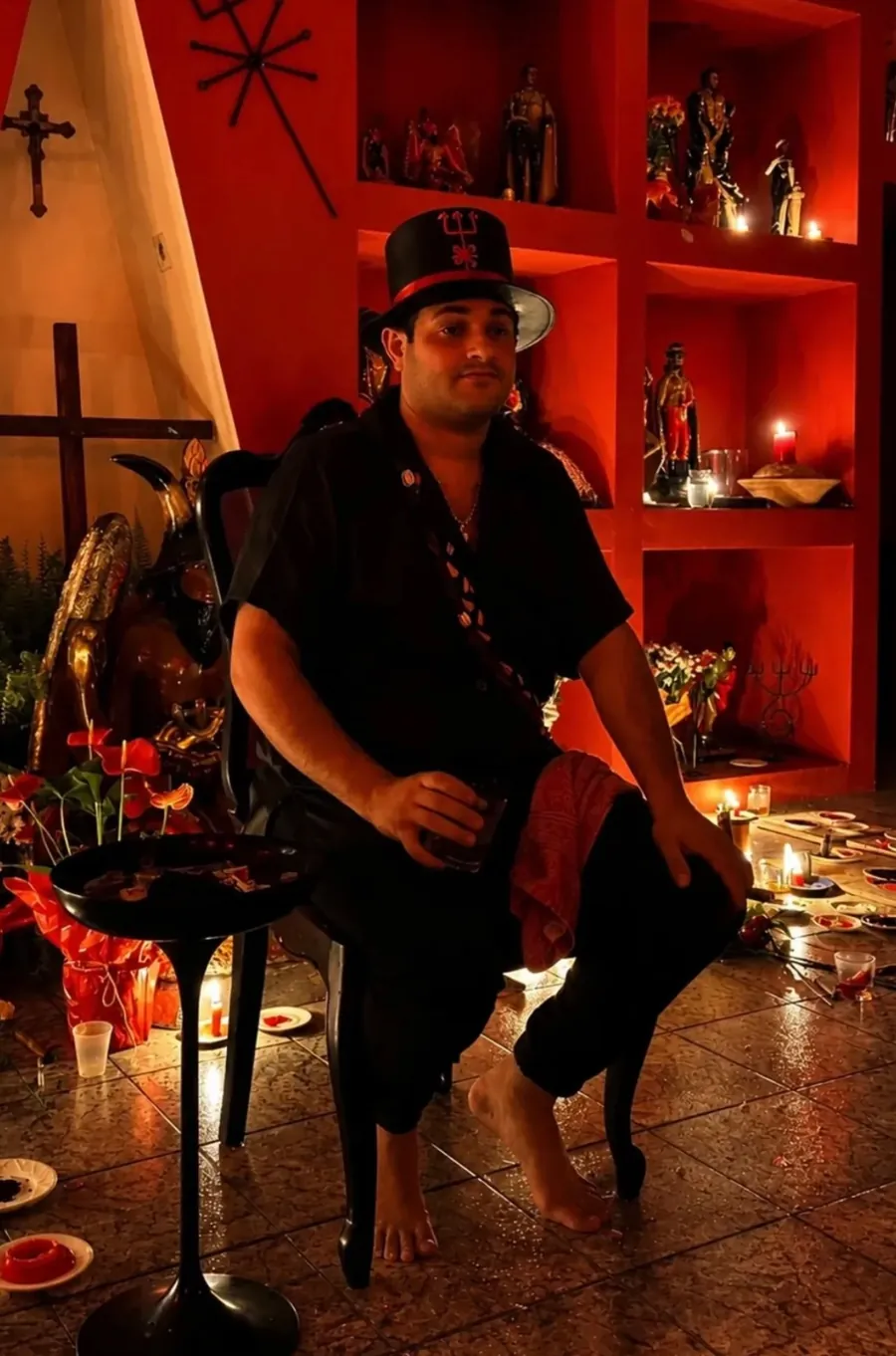 Pai Luiz Fernando de cartola preta com tridente, sentado na sala vermelha da Kimbanda, entre velas acesas e imagens do povo de Exu