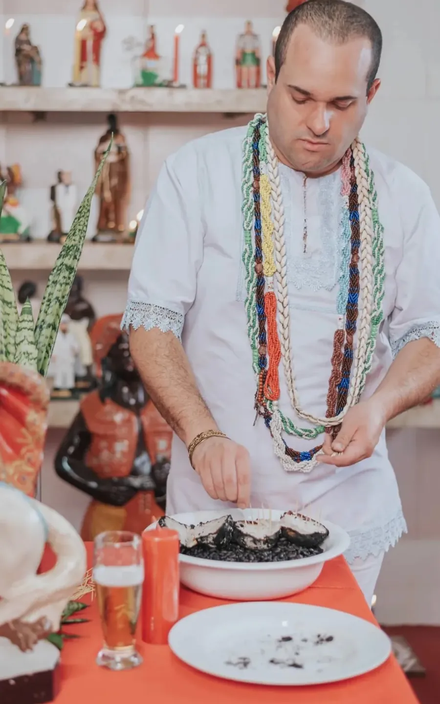 Pai Luiz Fernando de branco e guias preparando um trabalho em uma bacia, sobre a mesa vermelha com vela e imagens de santos ao fundo